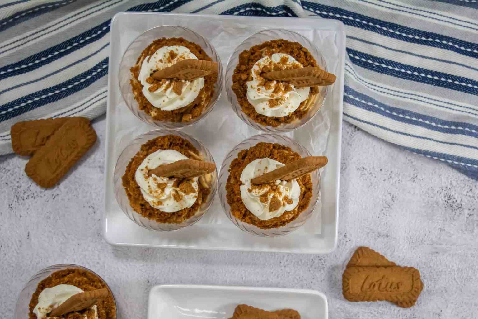 Four dessert cups topped with whipped cream and Lotus Biscoff cookies are arranged on a white tray, with extra cookies scattered nearby on a light surface.