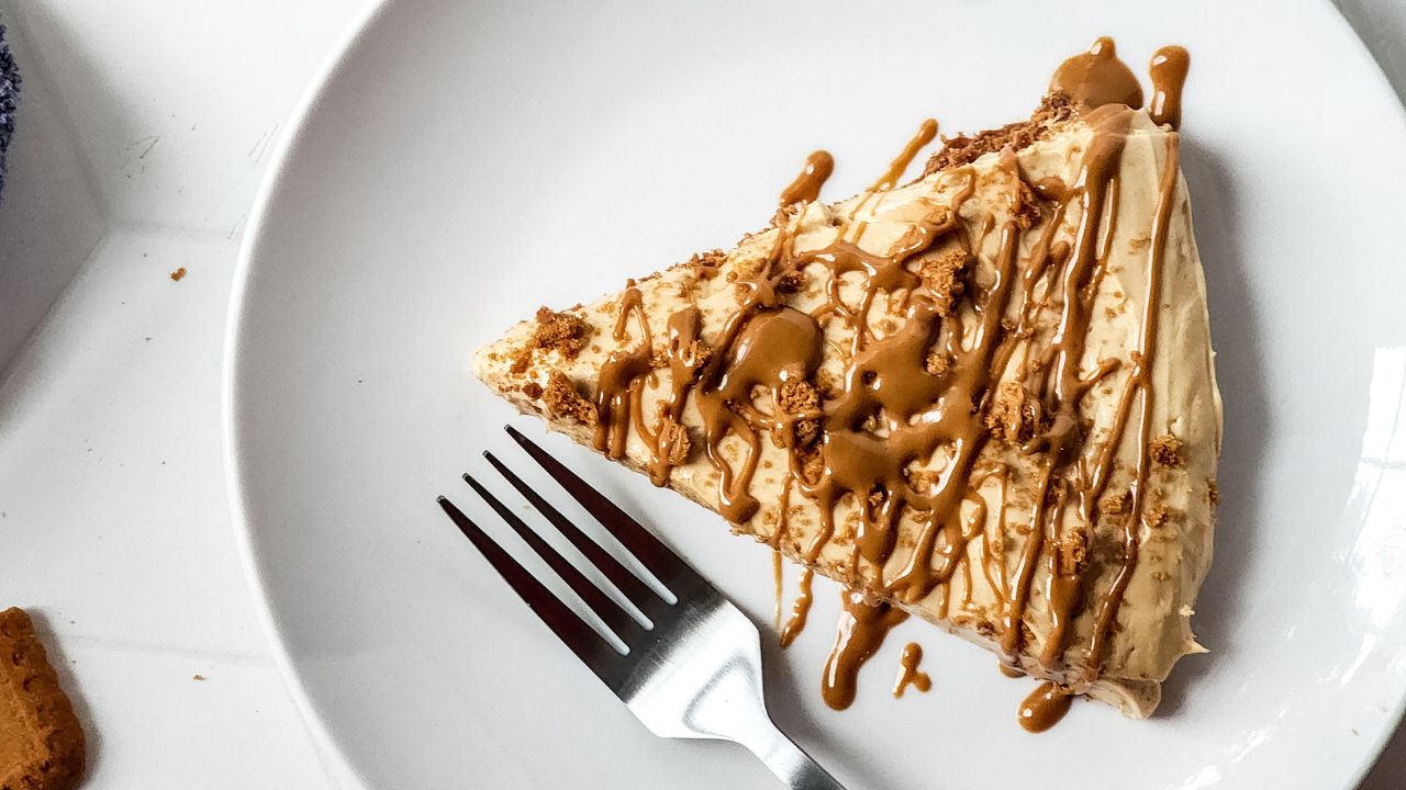 A slice of cake with light brown frosting and caramel drizzle on a white plate, accompanied by a fork.