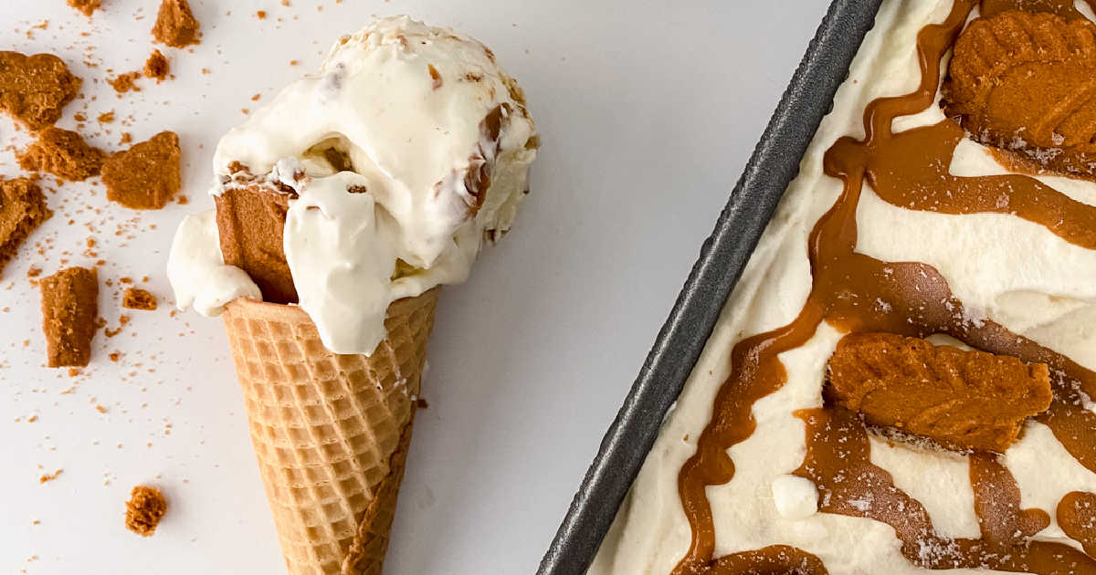 A scoop of vanilla ice cream with cookie pieces in a waffle cone next to a pan of ice cream topped with caramel and cookies on a white surface.