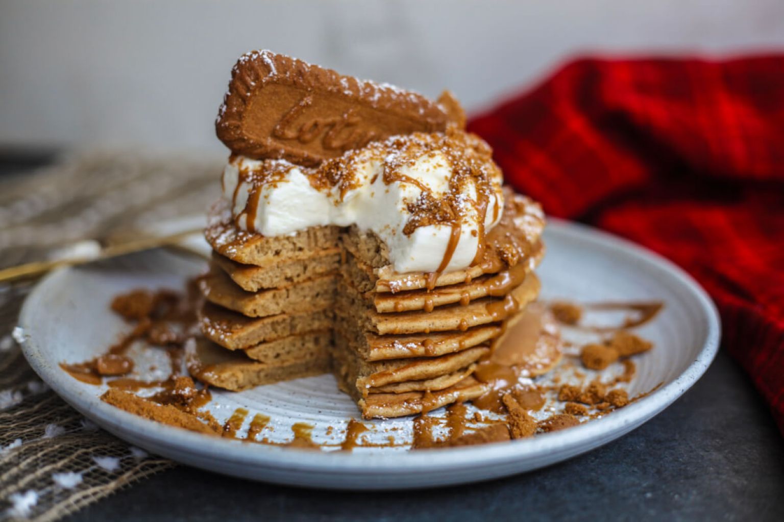 A stack of pancakes topped with whipped cream, a biscuit, and drizzled sauce sits on a plate with a piece cut out; a red cloth is in the background.