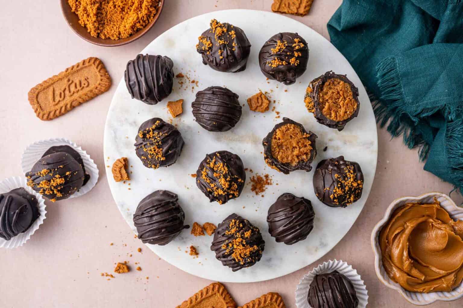 A marble plate with chocolate-covered truffles topped with cookie crumbs, surrounded by whole and broken cookies, a bowl of cookie butter, and a dark green cloth.