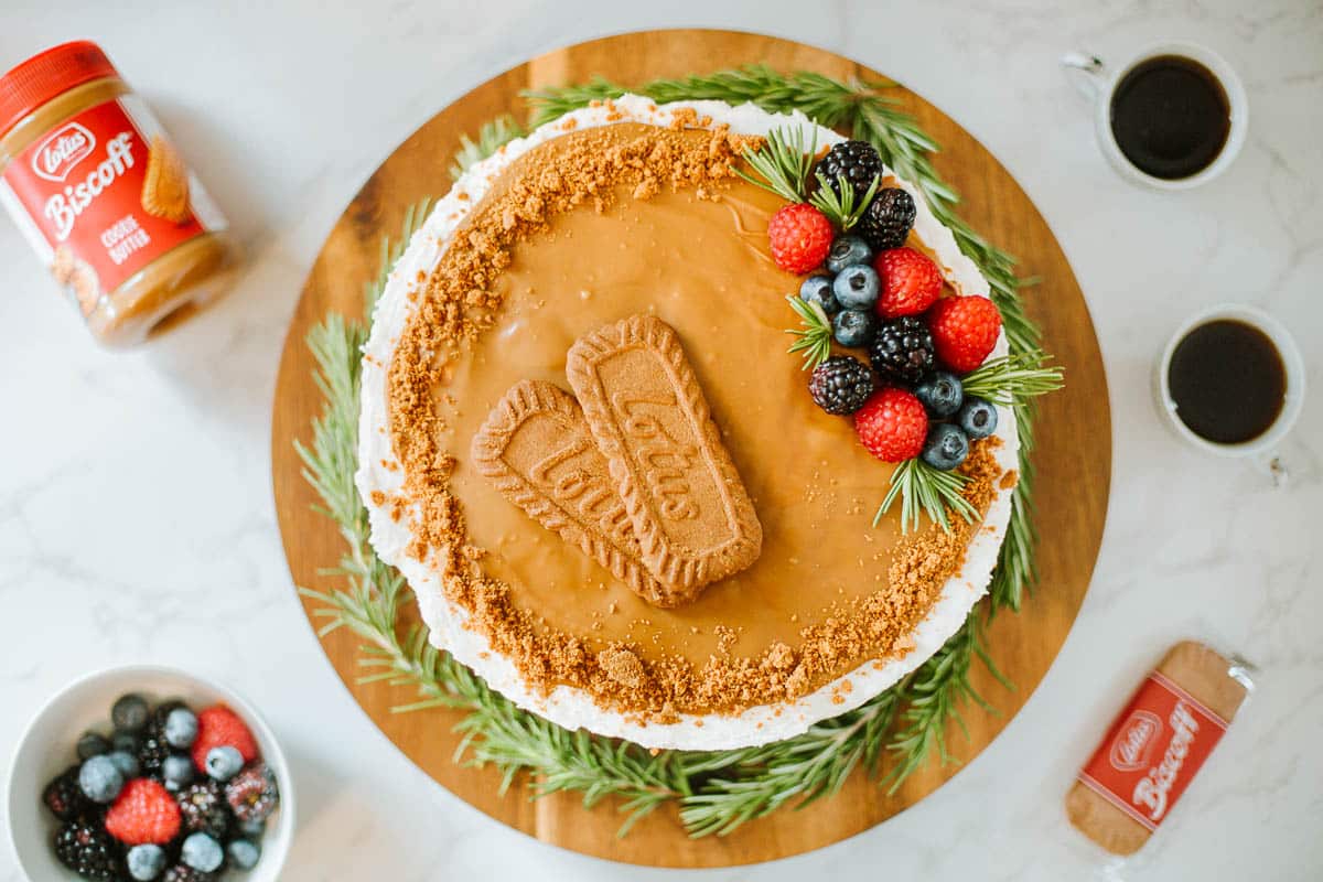 A round cake topped with Biscoff cookies, berries, and rosemary sits on a wooden board. Two cups of coffee, a bowl of berries, and Biscoff cookies are nearby.