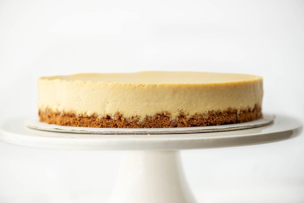 A plain cheesecake with a graham cracker crust sits on a white cake stand against a white background.