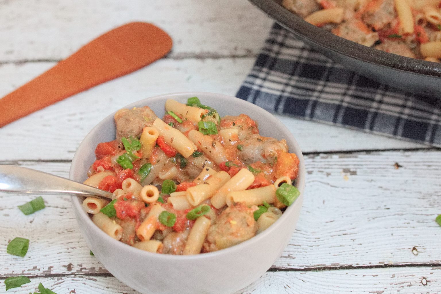 A white bowl filled with pasta, sausage pieces, diced tomatoes, and chopped green onions, with a spoon inside. A skillet and wooden spatula are in the background.