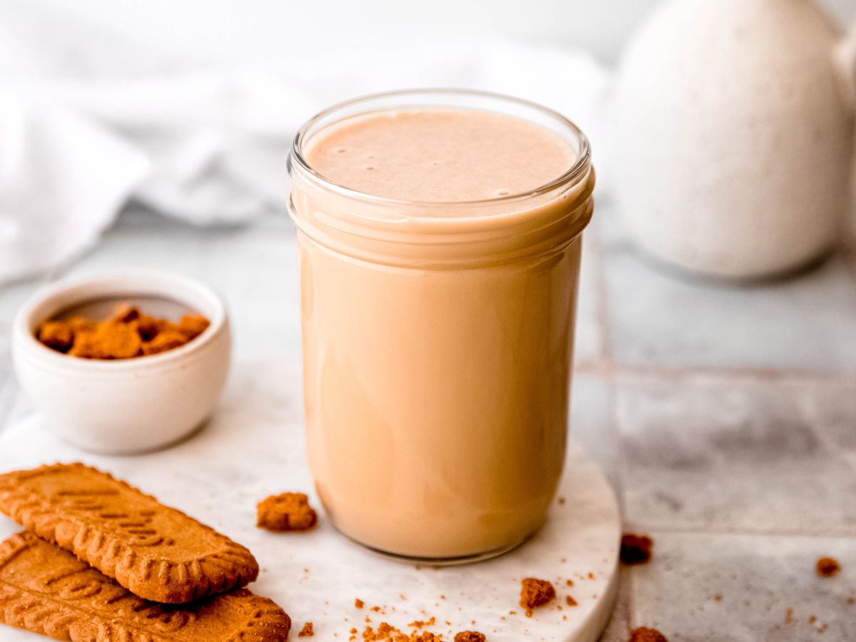 A glass jar filled with a light brown beverage sits on a round white surface, with crumbled cookies and whole cookies nearby.