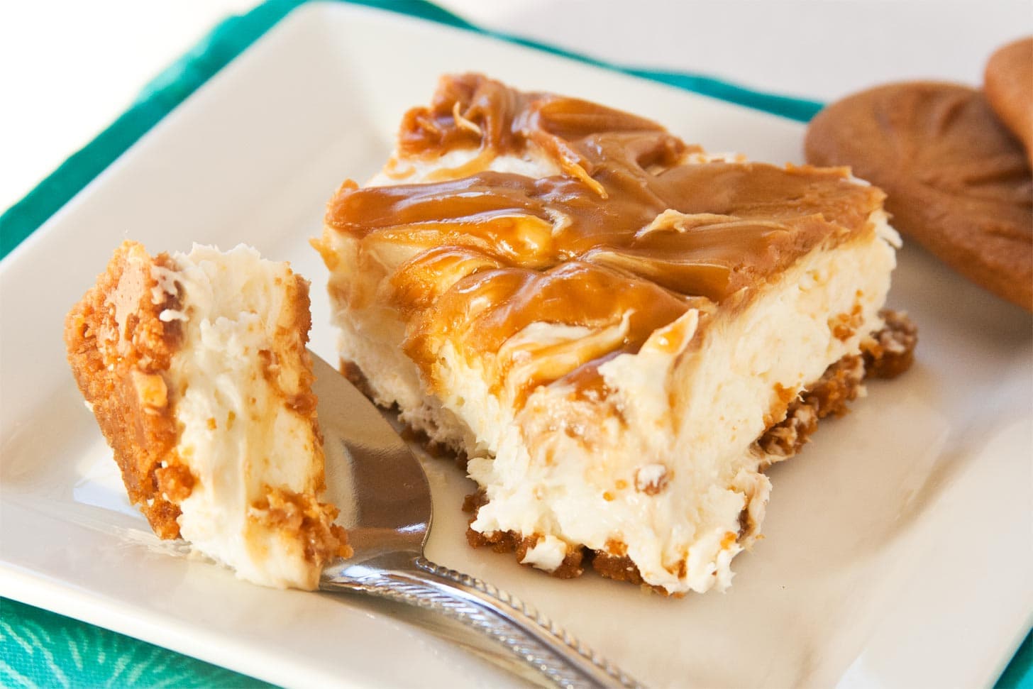 A slice of cheesecake with a caramel swirl topping and a crumbly crust sits on a white plate next to a fork holding a bite-sized piece.