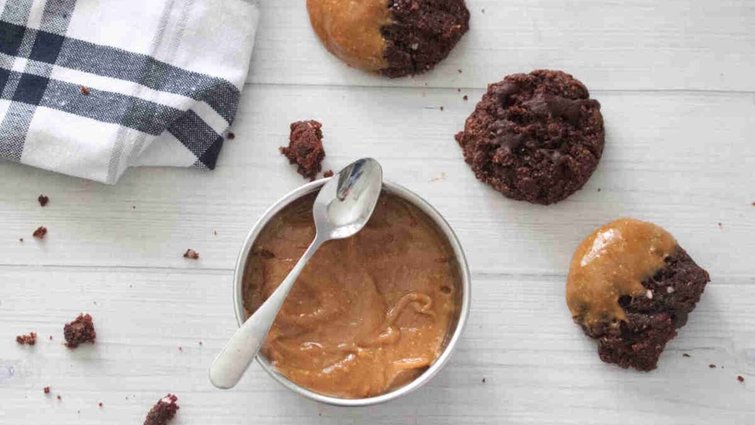 A bowl of peanut butter with a spoon, surrounded by chocolate cookies and a blue-striped napkin on a white surface.