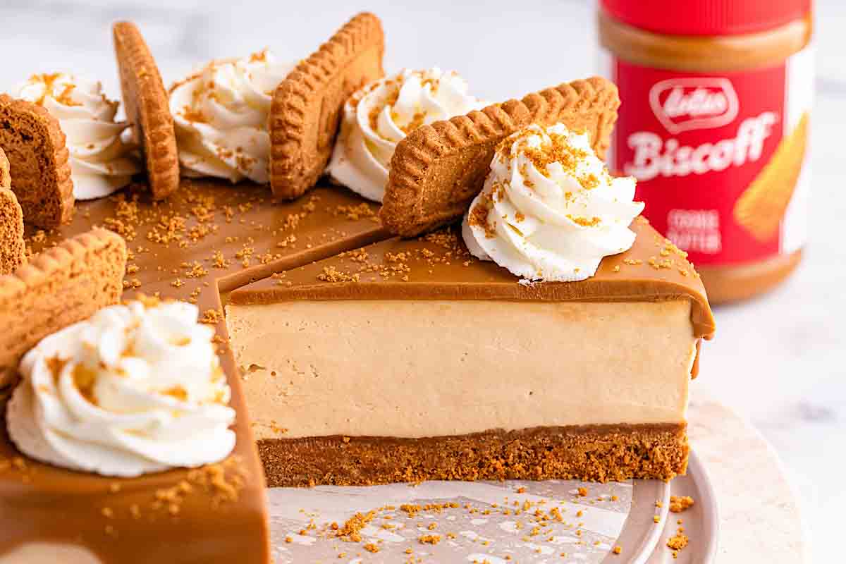 A sliced Biscoff cheesecake topped with whipped cream, Biscoff cookies, and cookie crumbs sits next to a jar of Biscoff spread.