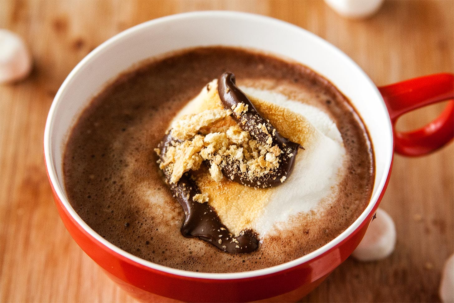 A red mug filled with hot chocolate topped with toasted marshmallow, melted chocolate, and crumbled graham crackers, placed on a wooden surface.