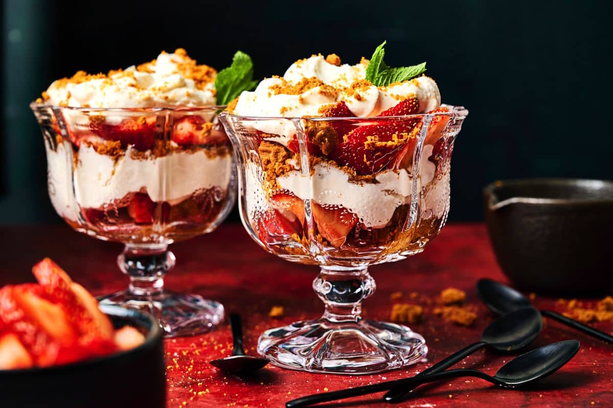Two glass dessert cups filled with layers of strawberries, whipped cream, and crumbled cookies, garnished with mint leaves, with spoons and a bowl of strawberries nearby.