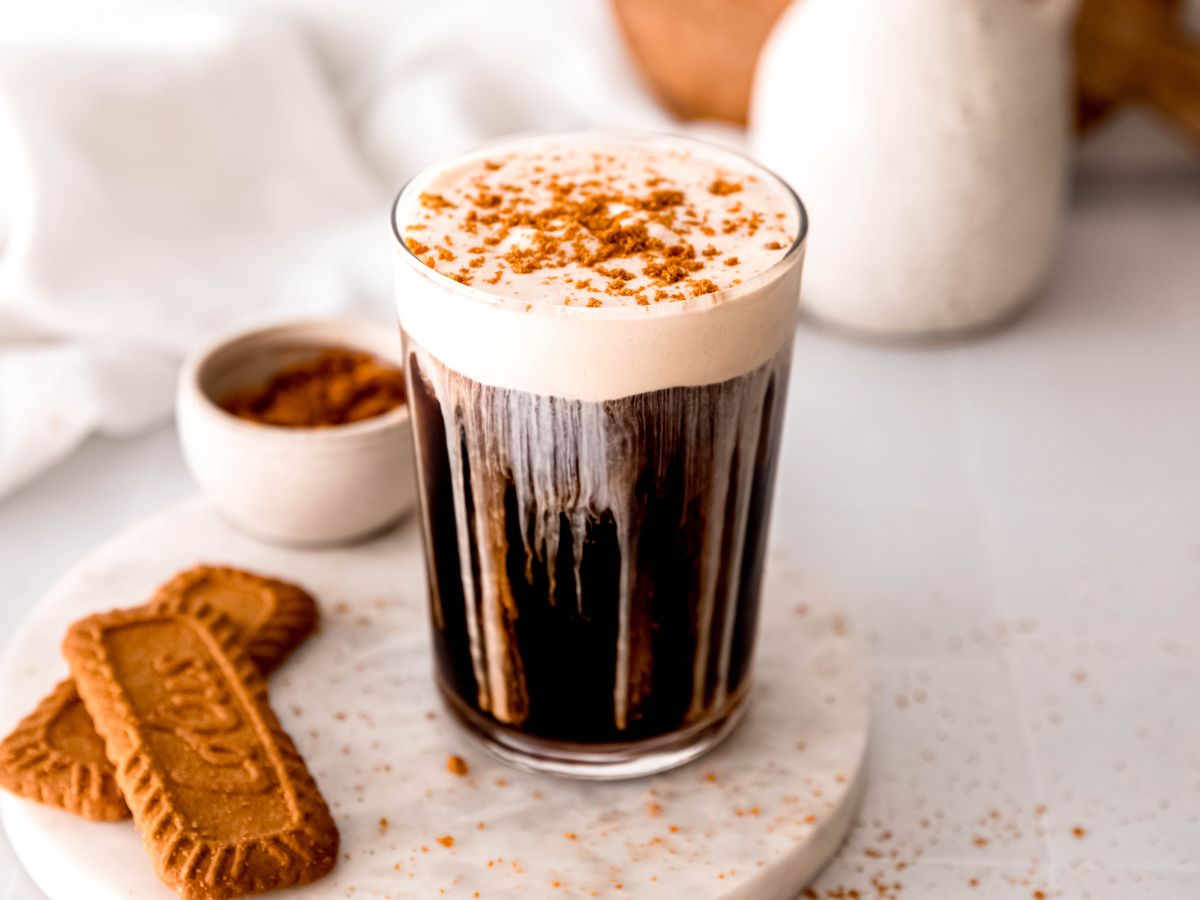 A glass of iced coffee topped with foam and cinnamon sits on a white surface beside two Biscoff cookies and a small bowl of cookie crumbs.