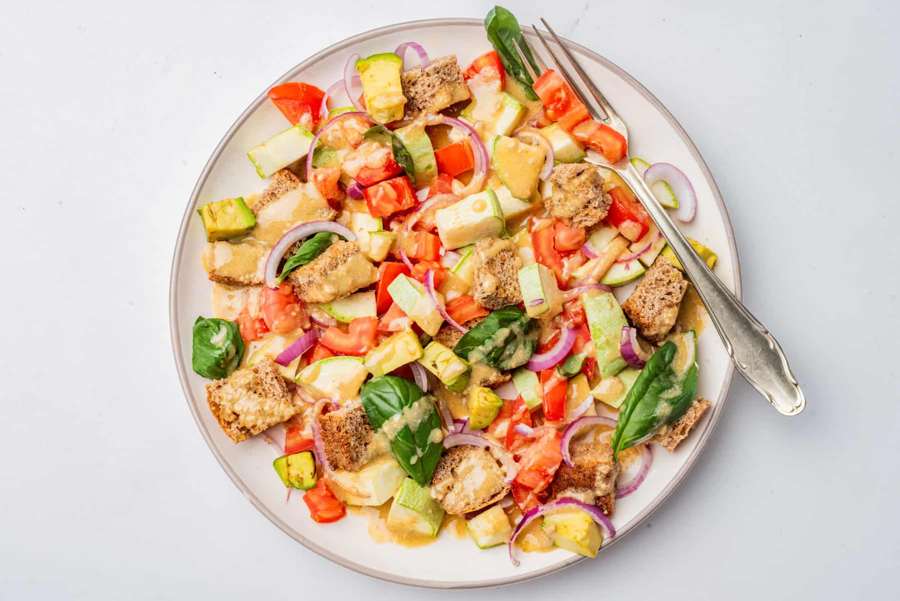 A plate of salad with diced tomatoes, avocado, red onion, croutons, spinach leaves, and creamy dressing. A fork and knife rest on the plate.