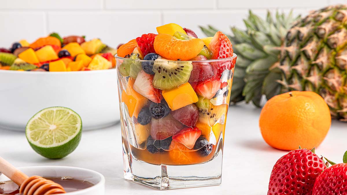 A glass filled with mixed fruit salad, including kiwi, strawberries, blueberries, grapes, and mango, sits on a table with fresh fruit and a bowl of more fruit salad in the background.
