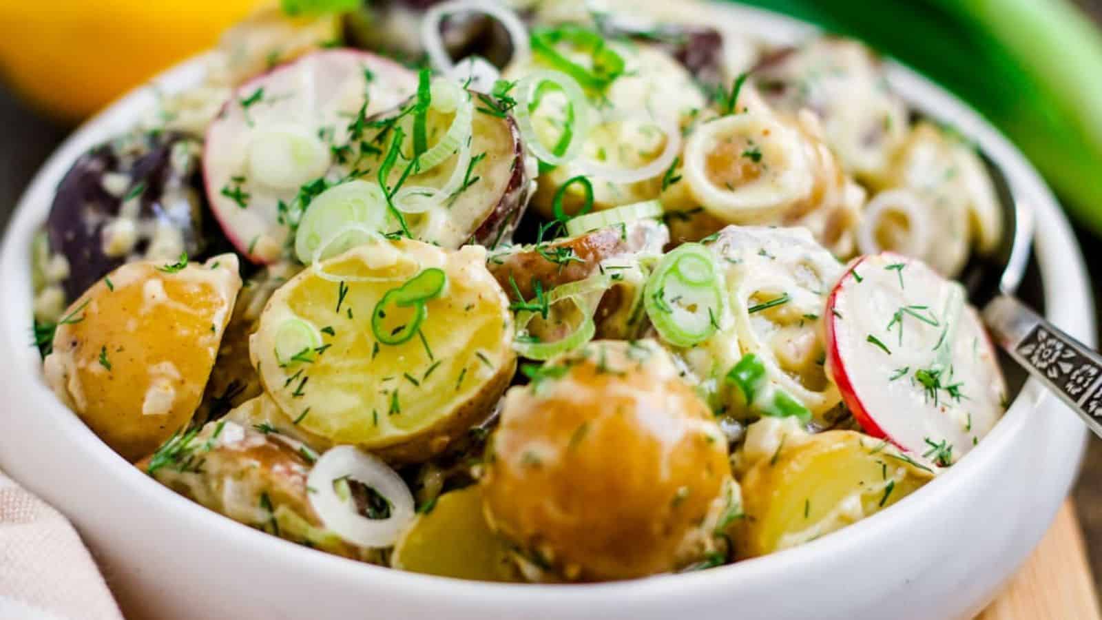 A bowl of creamy potato salad with sliced radishes, fresh dill, and chopped green onions, served with a spoon.
