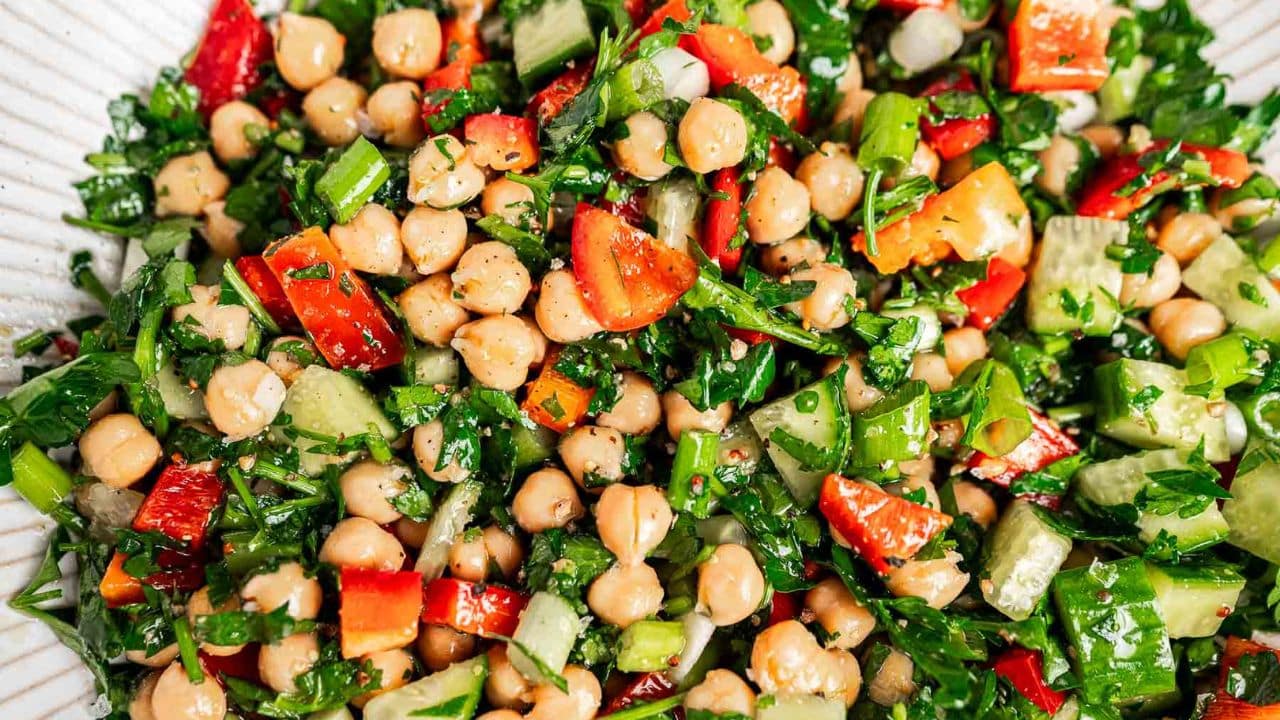 A close-up of a chickpea salad with chopped red bell peppers, cucumber, parsley, and green onions mixed together.