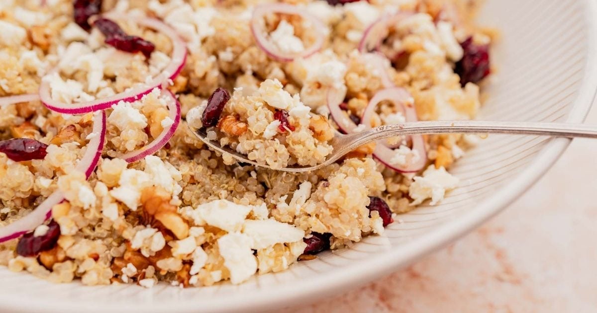 A close-up of a quinoa salad with feta cheese, sliced red onions, walnuts, and dried cranberries in a bowl with a spoon.