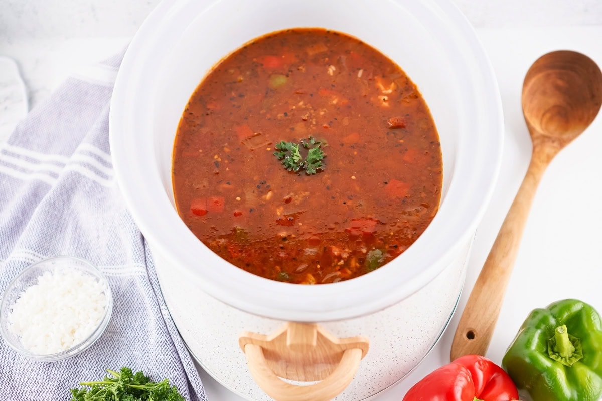A white slow cooker filled with tomato-based soup, garnished with parsley, sits on a countertop near a wooden spoon, green and red bell peppers, and a bowl of grated cheese.