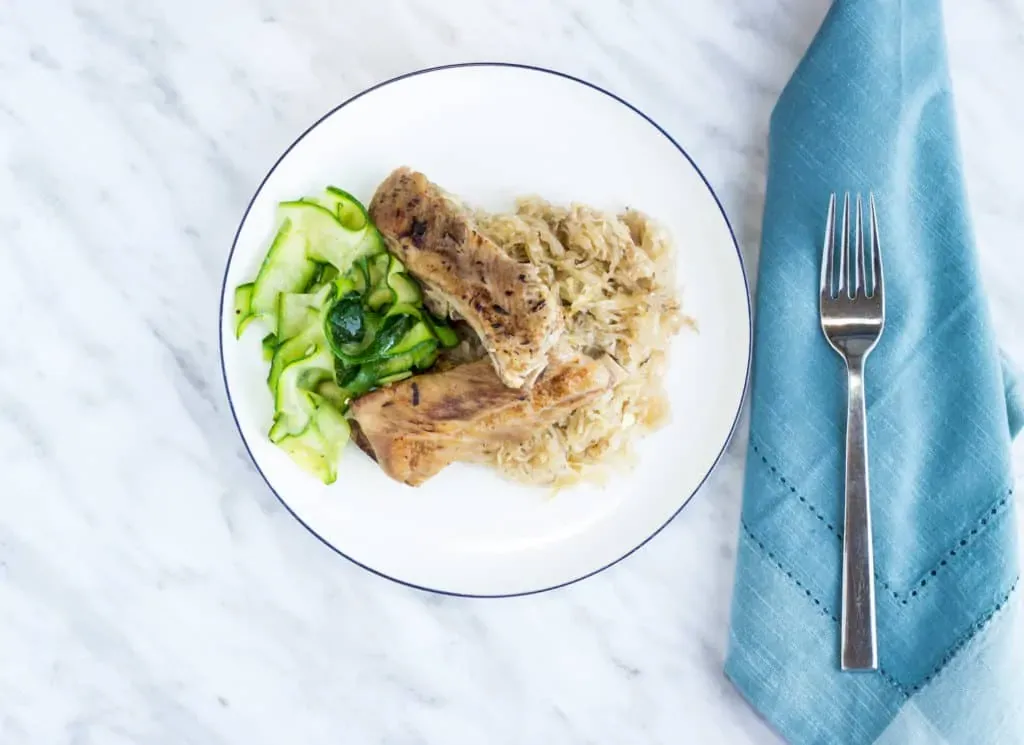 A white plate with braised pork ribs, sauerkraut, and zucchini ribbons on a marble surface next to a blue napkin and fork.