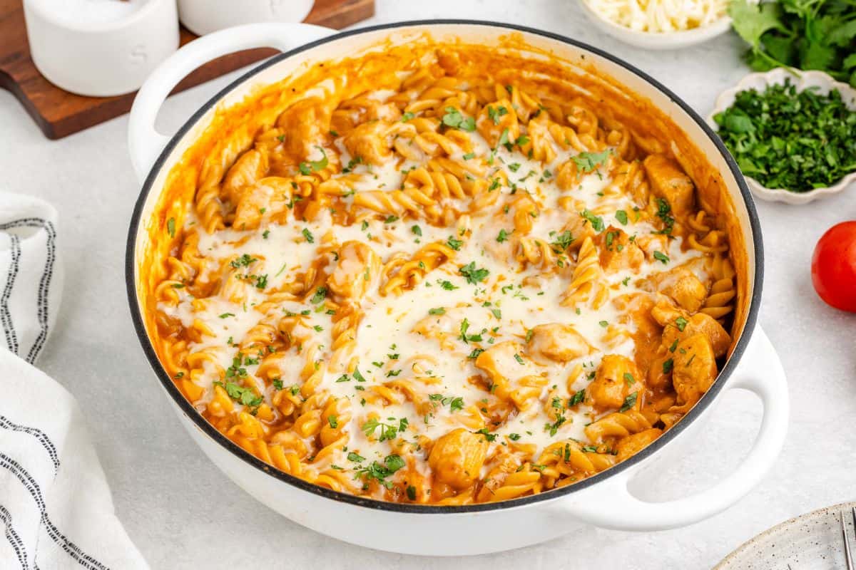 A white pot filled with rotini pasta, chicken pieces, tomato sauce, and melted cheese, garnished with chopped parsley.