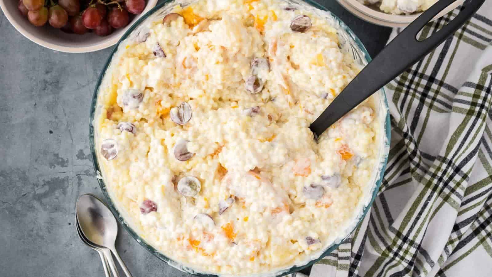 A bowl of creamy ambrosia salad with grapes and mandarin oranges, next to a plaid napkin, metal spoons, and a small bowl of grapes.