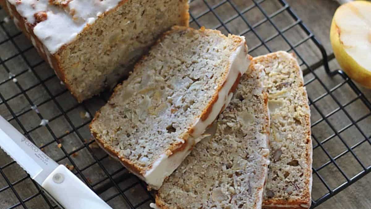 Sliced loaf of glazed bread with visible apple and nut pieces on a cooling rack, next to a knife and a partially visible apple.