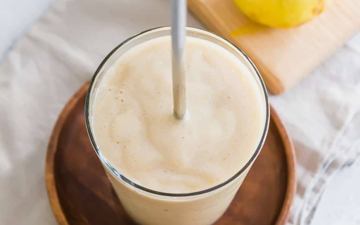 A glass of creamy banana smoothie with a straw, placed on a wooden tray, with a blurred lemon on a cutting board in the background.