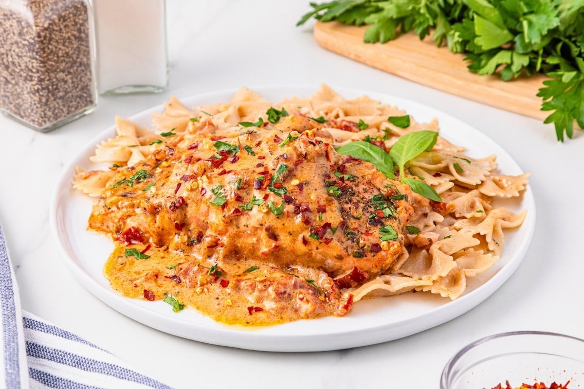 A plate of creamy chicken in a seasoned sauce served over bowtie pasta, garnished with herbs, with parsley and seasonings in the background.