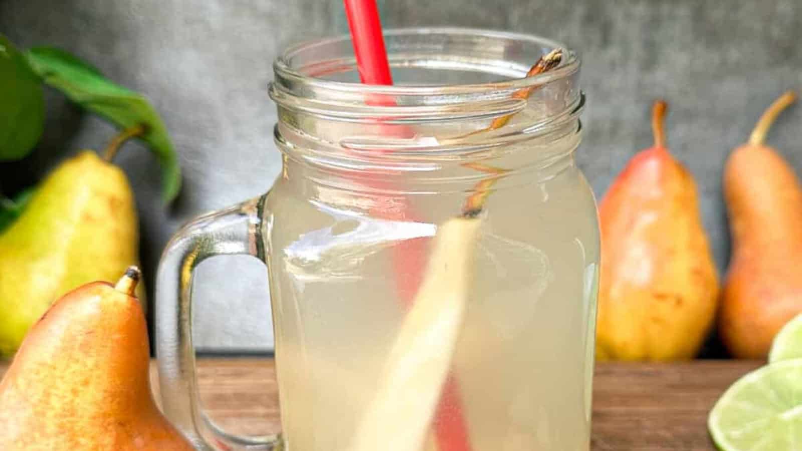 A mason jar filled with light-colored pear juice, a red straw, and a slice of pear, with whole pears and lime slices in the background.