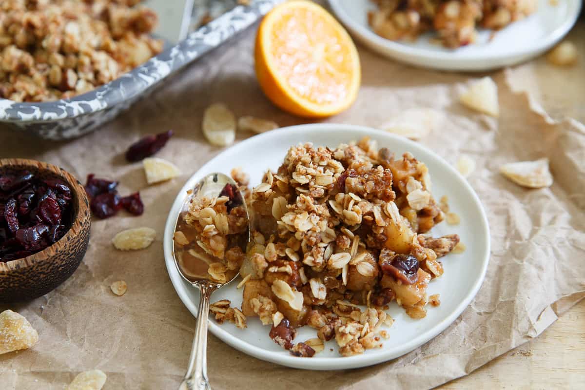 A plate with a serving of fruit crumble topped with oats, a spoon, dried cranberries, and a halved orange in the background.