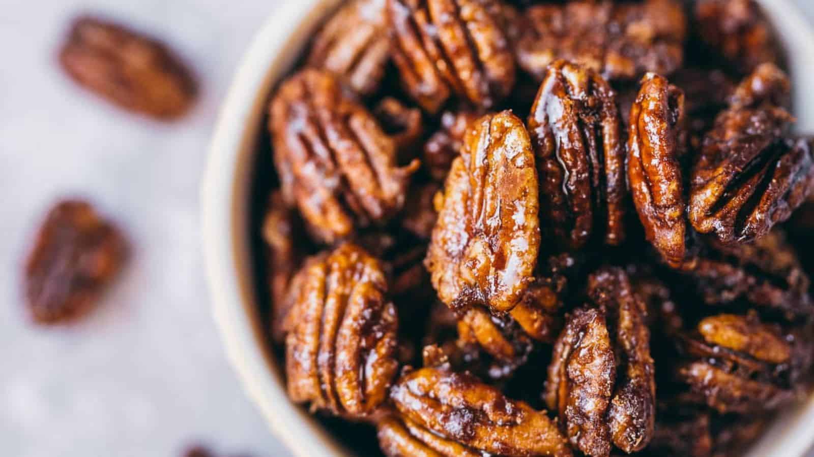 A close-up view of glazed pecans in a bowl, with a few pieces scattered outside the bowl on a light surface.
