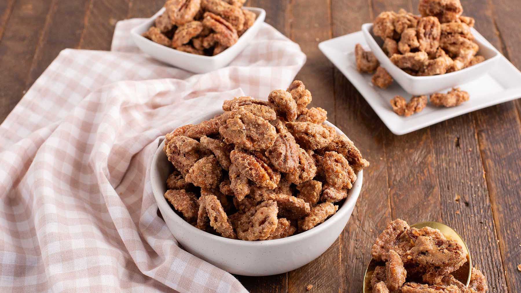 Bowls of candied pecans are arranged on a wooden table with a checked napkin.