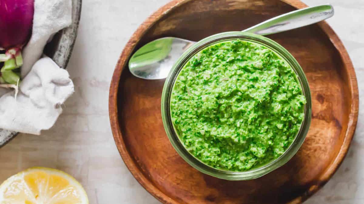 A glass jar filled with green pesto sits on a wooden plate with a metal spoon beside it. Part of a lemon and a bowl of vegetables are partially visible nearby.