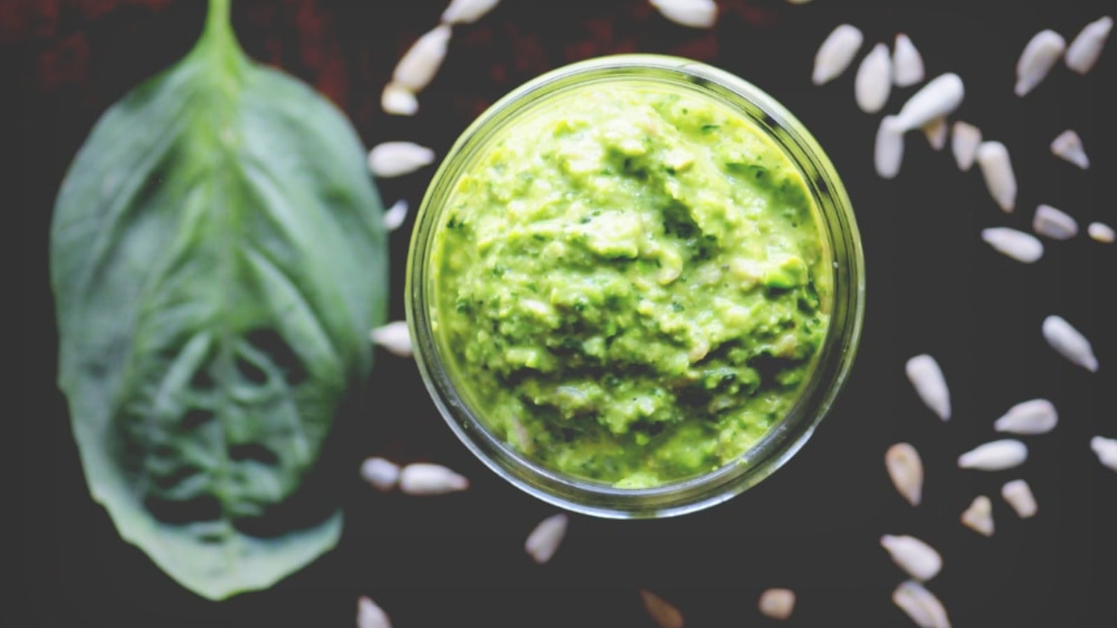 A glass jar of green pesto sauce is shown beside a basil leaf and scattered sunflower seeds on a dark surface.