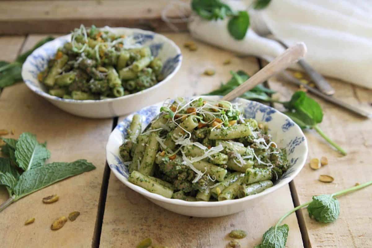 Two bowls of pasta with green pesto sauce, topped with grated cheese and herbs, placed on a rustic wooden table with mint leaves and seeds scattered around.