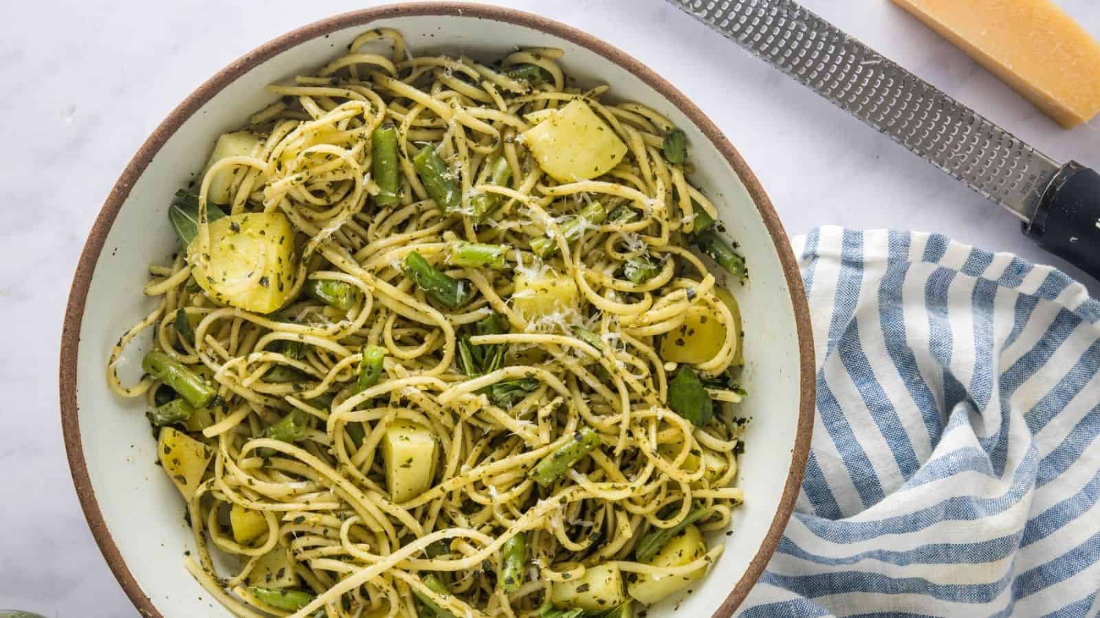 A bowl of spaghetti with green beans, potato pieces, and pesto sauce, placed on a white surface next to a striped cloth, a cheese grater, and a block of cheese.