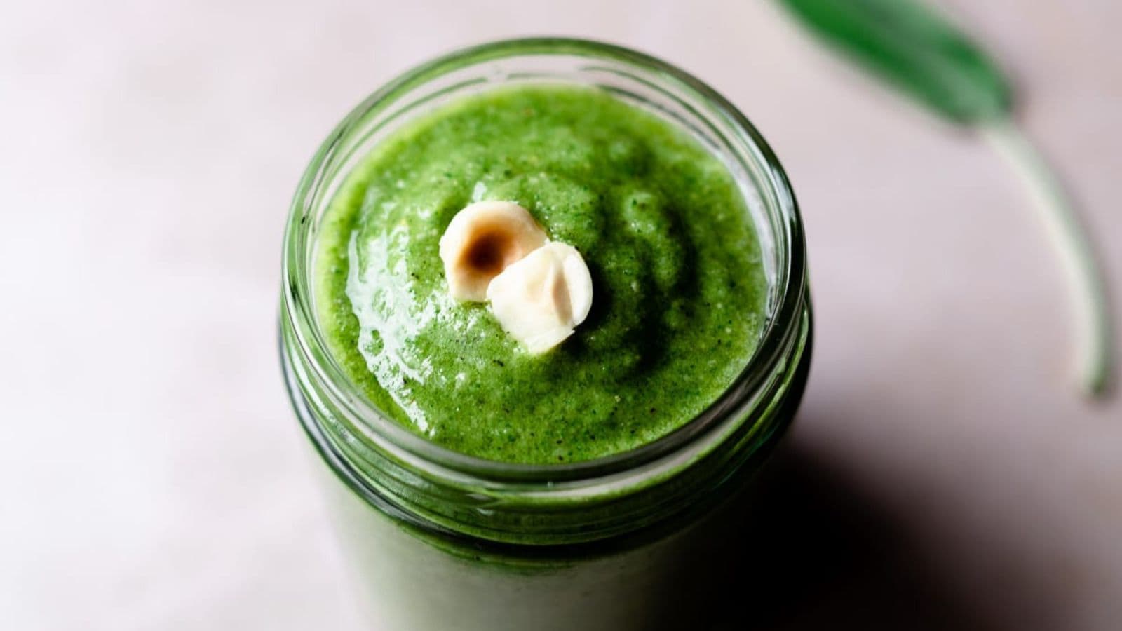 A glass jar filled with thick green smoothie, topped with two hazelnuts, sits on a light surface with a green leaf in the background.