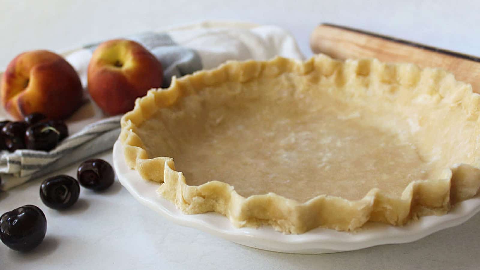 Unbaked pie crust in a white dish sits on a counter with peaches, cherries, a kitchen towel, and a rolling pin nearby.