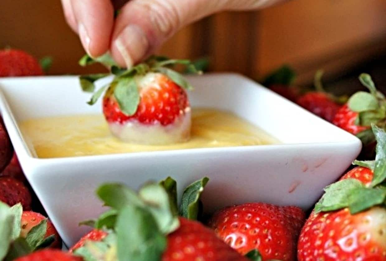 A hand dips a fresh strawberry into a bowl of creamy yellow sauce, with more strawberries surrounding the bowl.