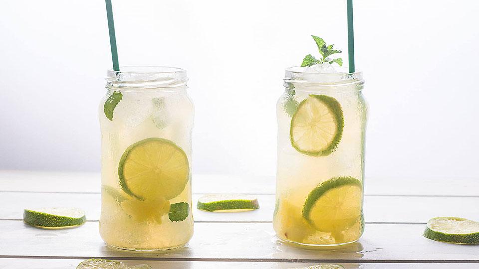 Two mason jars filled with iced lemonade, lime slices, mint leaves, and green straws sit on a white wooden surface with scattered lime slices.