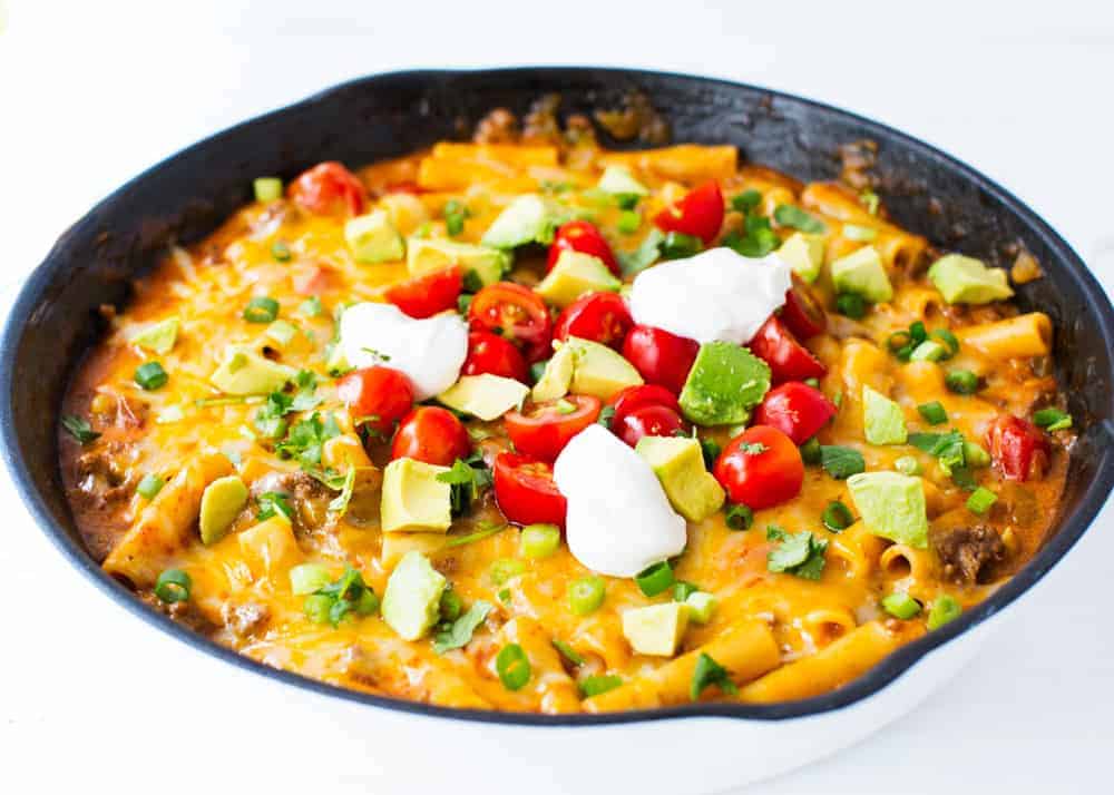 A skillet filled with cheesy pasta topped with chopped avocado, cherry tomatoes, green onions, and dollops of sour cream.