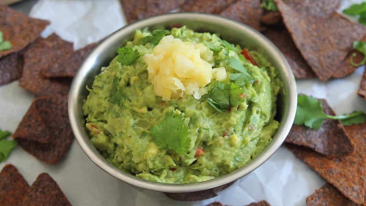 A bowl of guacamole topped with a small amount of chopped pineapple, garnished with cilantro, surrounded by brown tortilla chips.