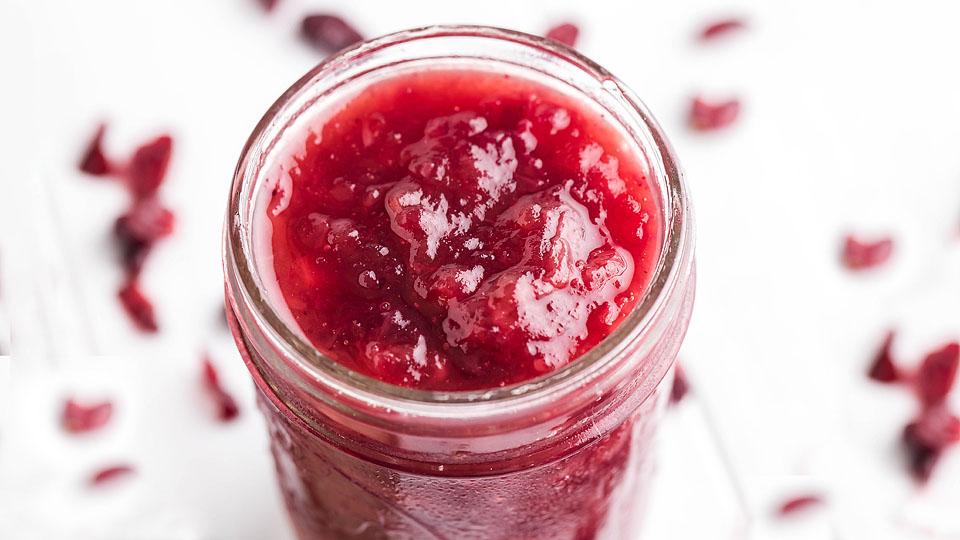 A glass jar filled with chunky red cranberry sauce, seen from above, with scattered cranberries in the background.