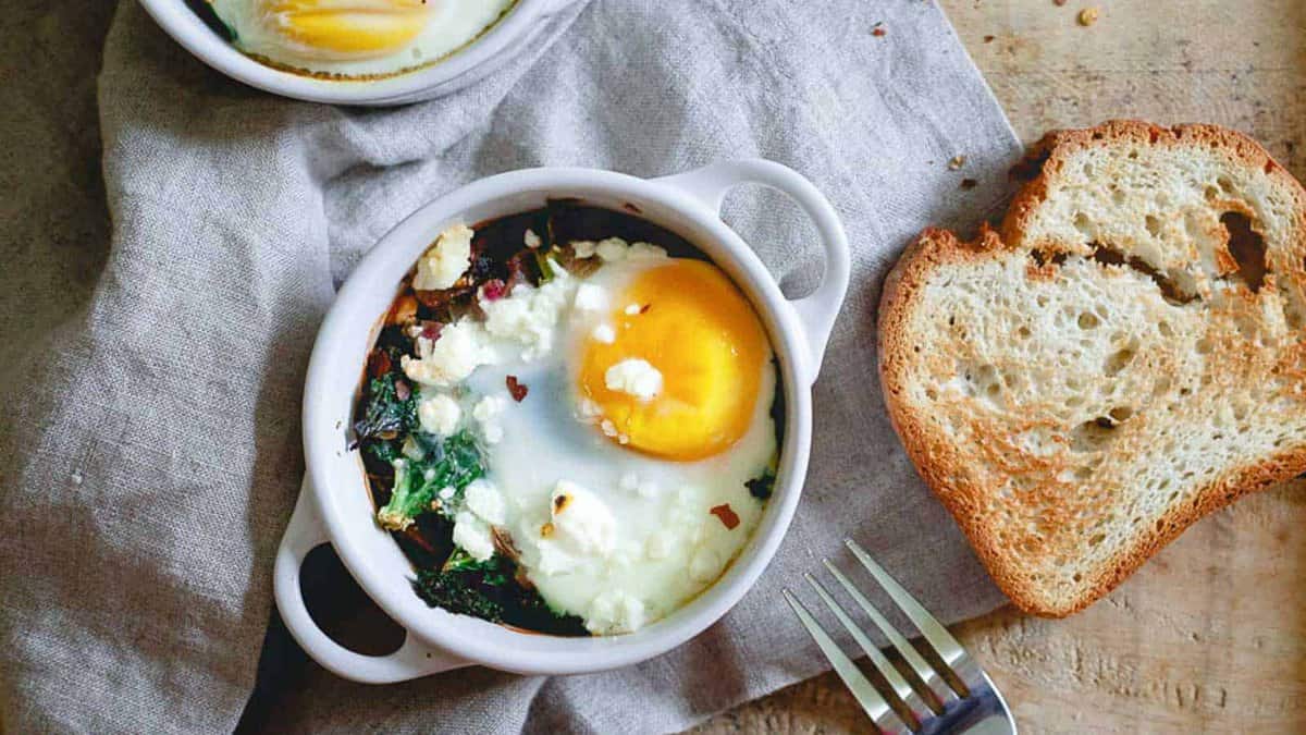 A baked egg with greens and cheese in a small white dish, served with two slices of toasted bread on the side and a fork placed nearby.