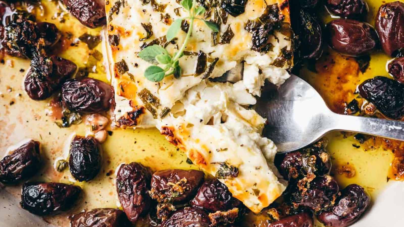 A fork holds a piece of baked feta cheese garnished with herbs, surrounded by roasted black olives and olive oil.
