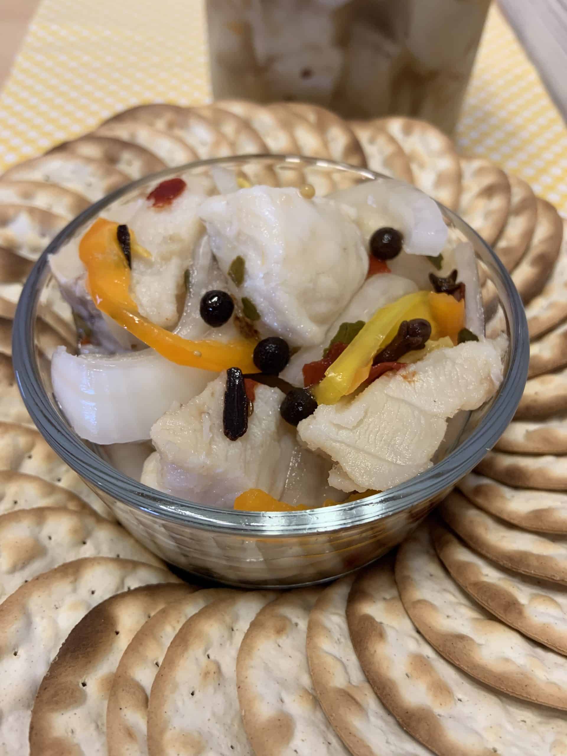 A glass bowl of pickled fish with onions, peppers, and spices is surrounded by a ring of round crackers on a plate.