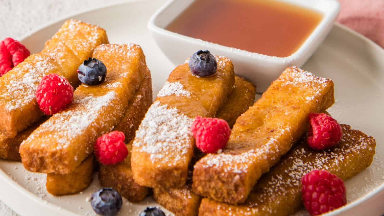 French toast sticks dusted with powdered sugar, garnished with raspberries and blueberries, served with a small bowl of syrup on a white plate.