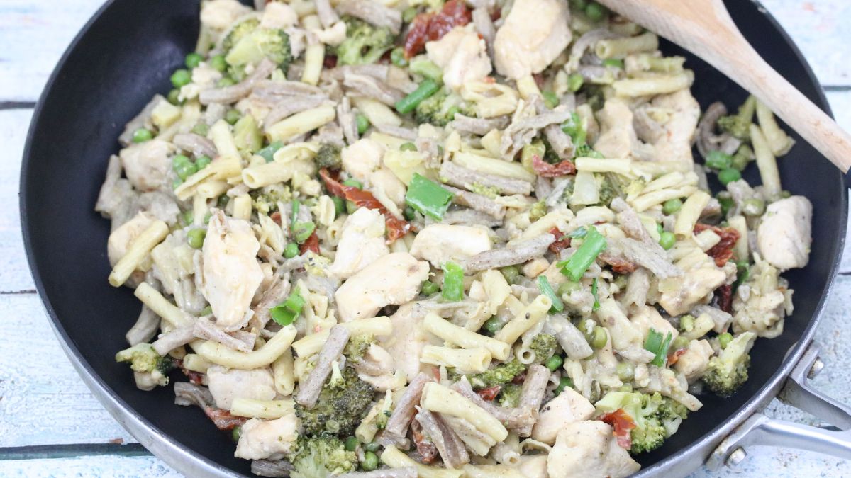 A skillet containing mixed pasta with chicken pieces, broccoli, sun-dried tomatoes, and green onions, stirred together with a creamy sauce.