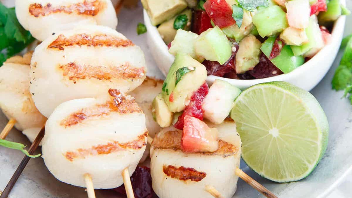 Three grilled scallops on skewers are served next to a bowl of diced avocado, cucumber, and strawberry salad, with a lime half on the side.
