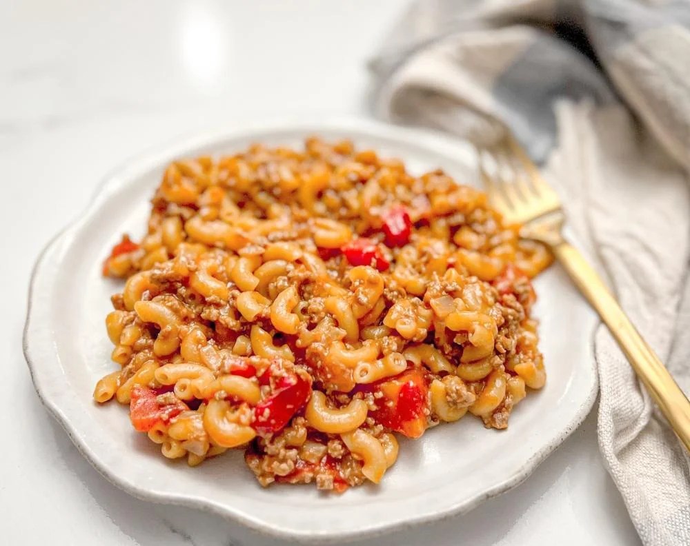 A plate of cooked macaroni pasta mixed with ground meat and diced red peppers, served on a white plate with a gold fork beside it.