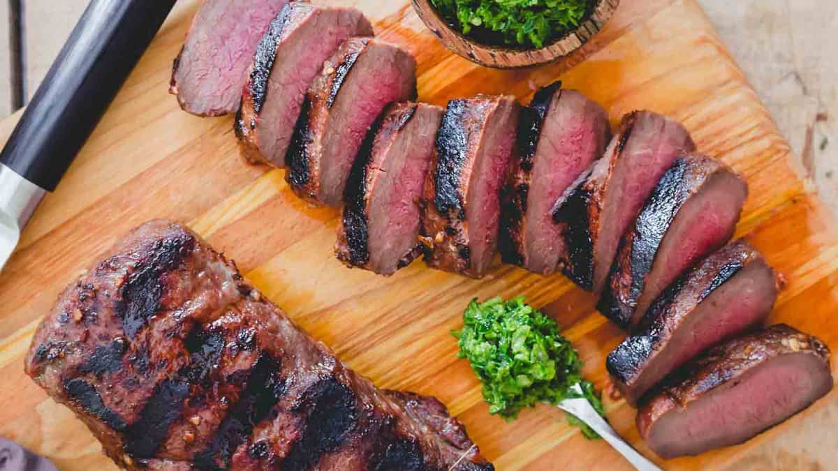 Sliced grilled steak with a charred exterior is arranged on a wooden cutting board with a small portion of green sauce on the side.